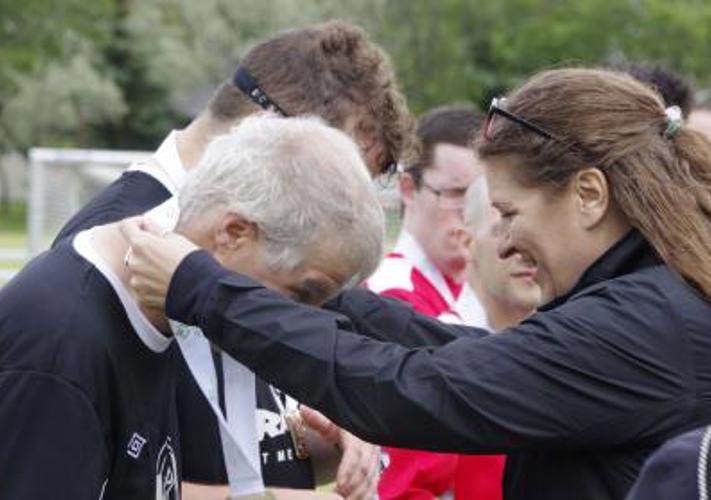 A woman on the right, wearing a black jacket, is placing a white medal around a male athlete's head. The woman is smiling and the man is bending forward so the medal could be placed around his neck.