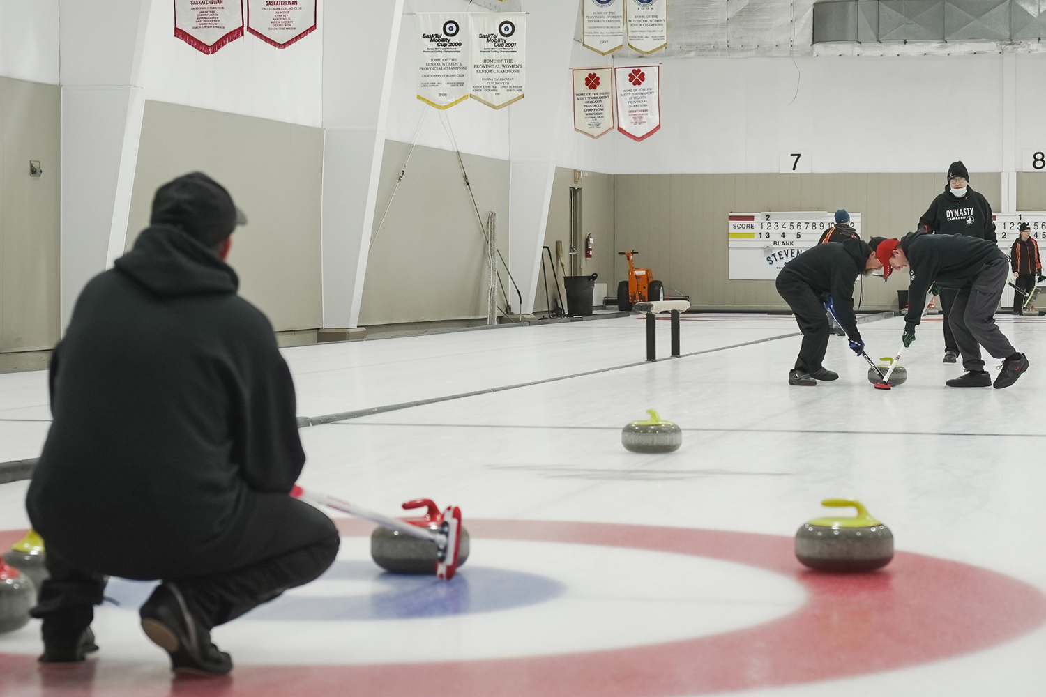 An action shot photo of a curling game. Three athletes are on the ice. One is crouching down next to the circles and the curling stones close to the camera. In the distance you can see two other athletes using their curling sticks to help the curling stone move across the ice.