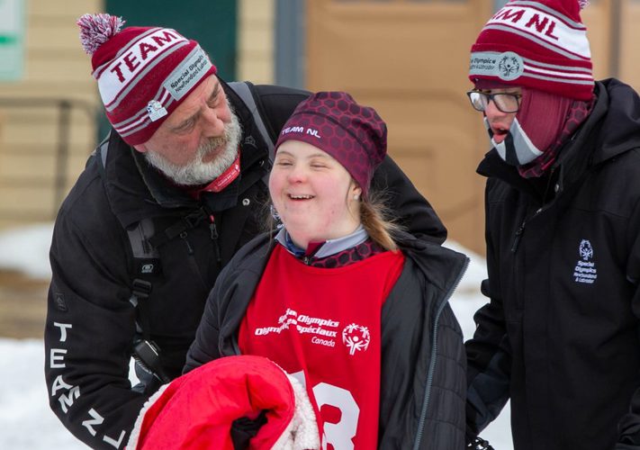 A photo of a volunteer and two athletes. They are all wearing Special Olympics Newfoundland & Labrador winter attire. The volunteer is standing behind the athletes looking around at them. The athletes are smiling.