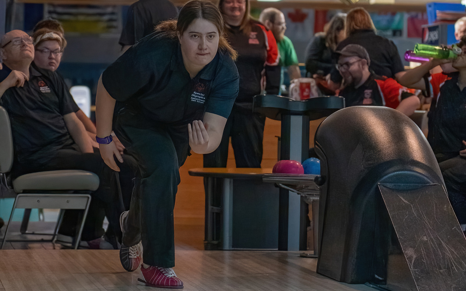An action shot photo of a bowling athlete. She is at the start of the lane, mid throw, and you can see the blue bowling ball start to roll down the aisle. Behind the athlete, you can see her teammates smiling and watching her turn.