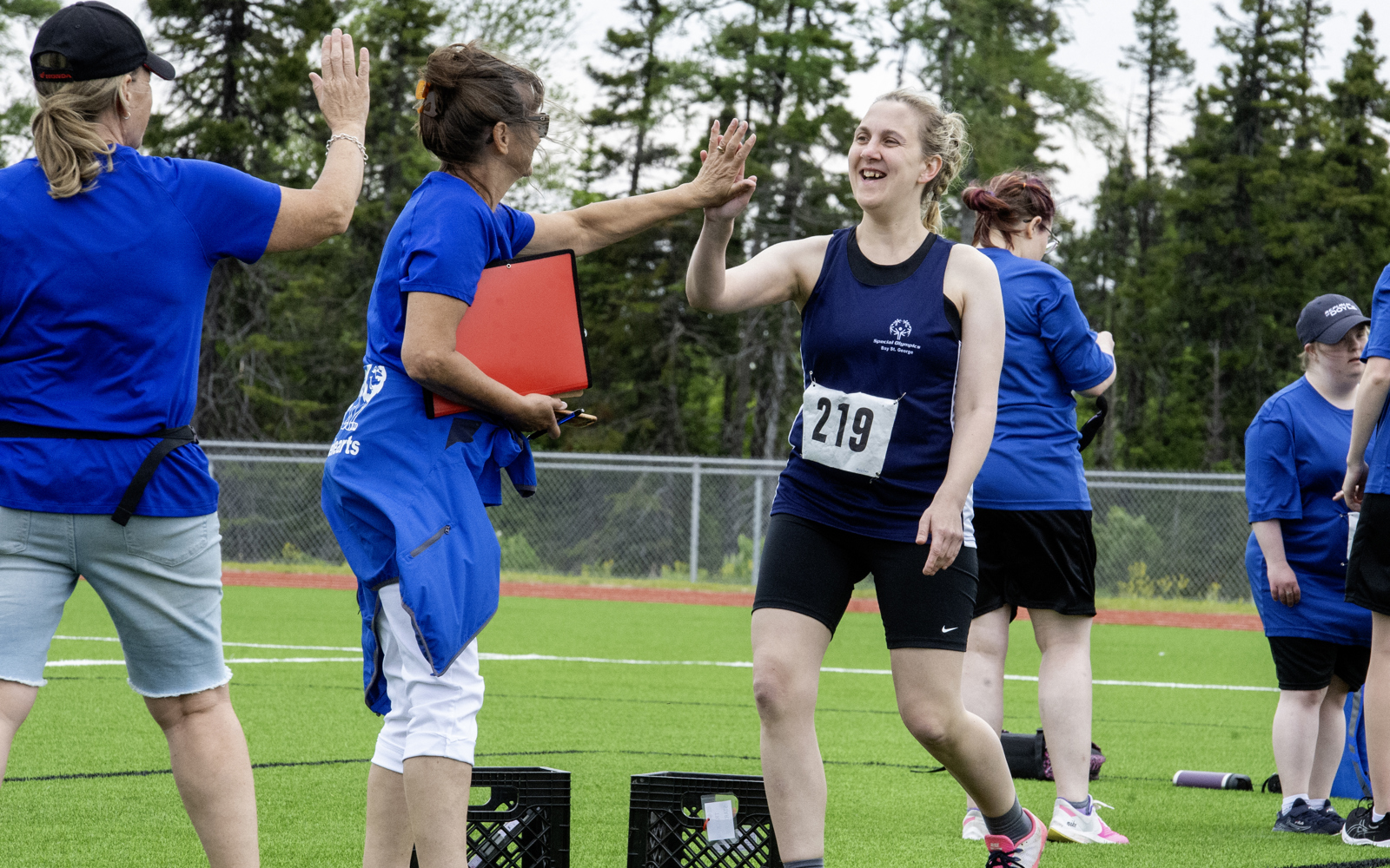 An athlete, on the sidelines after her run, is high-fiving with her coach. Her coach is holding a red binder and wearing a blue t-shirt with a red jacket wrapped around her waste. The athlete is wearing a blue tank top with black biker shorts. She has the number 219 pinned to her shirt.