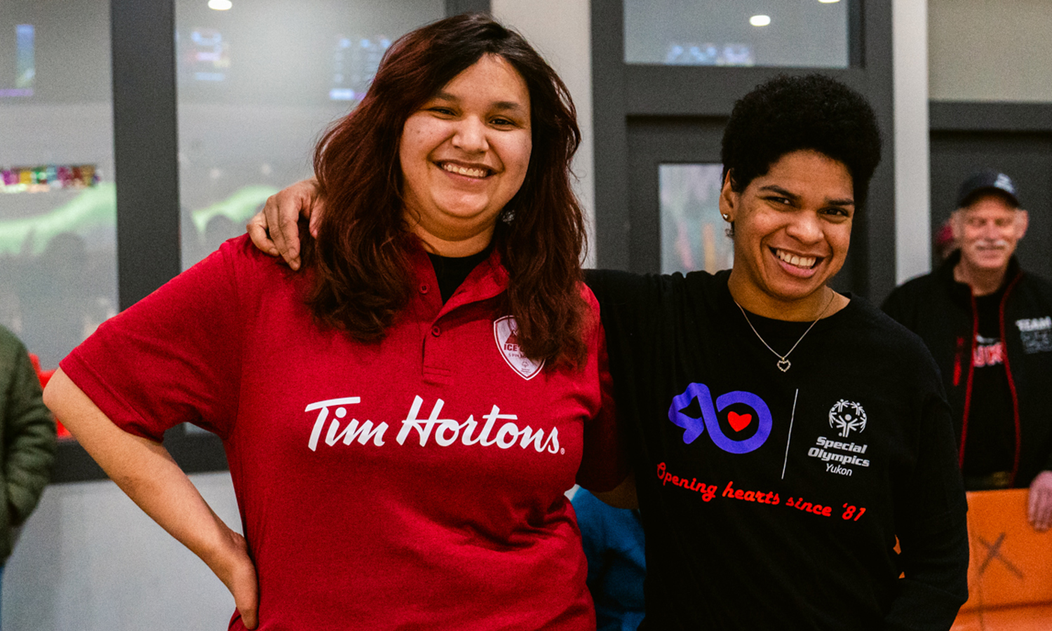 Two bowling players with their arms wrapped around behind each other, are smiling for the camera. The player on the left is wearing a red Tim Hortons collared shirt. The player on the right is wearing a black Special Olympics Yukon shirt.